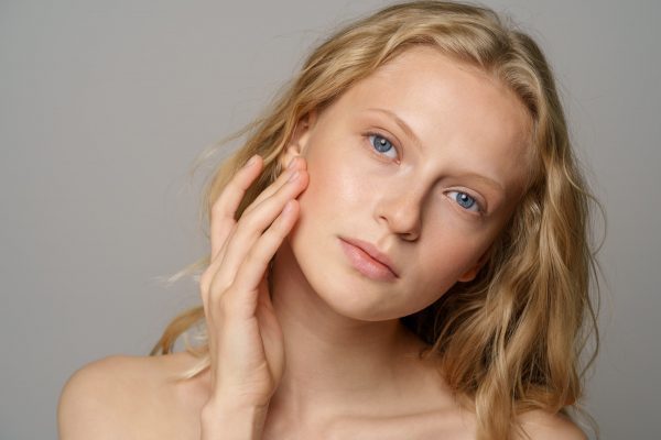 Young woman face with blue eyes, curly natural blonde hair, has no makeup, touching her soft skin